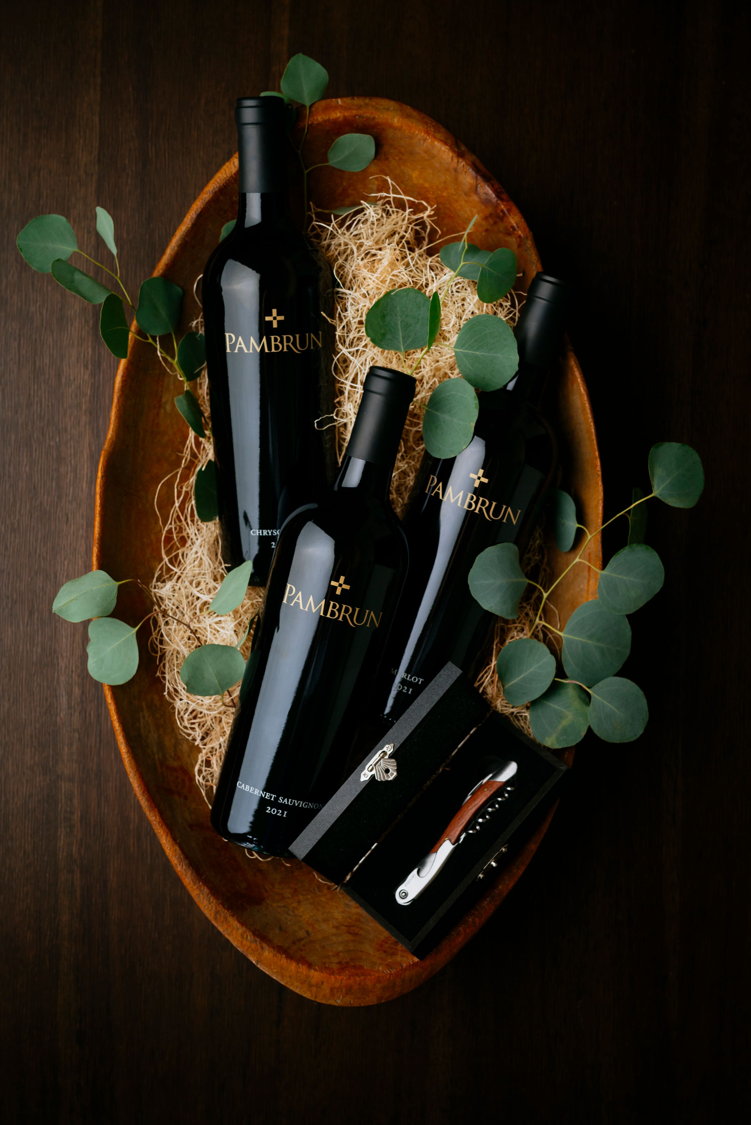 Black wine bottles nestled in straw with eucalyptus leaves in a rustic wooden serving bowl on dark wood surface.