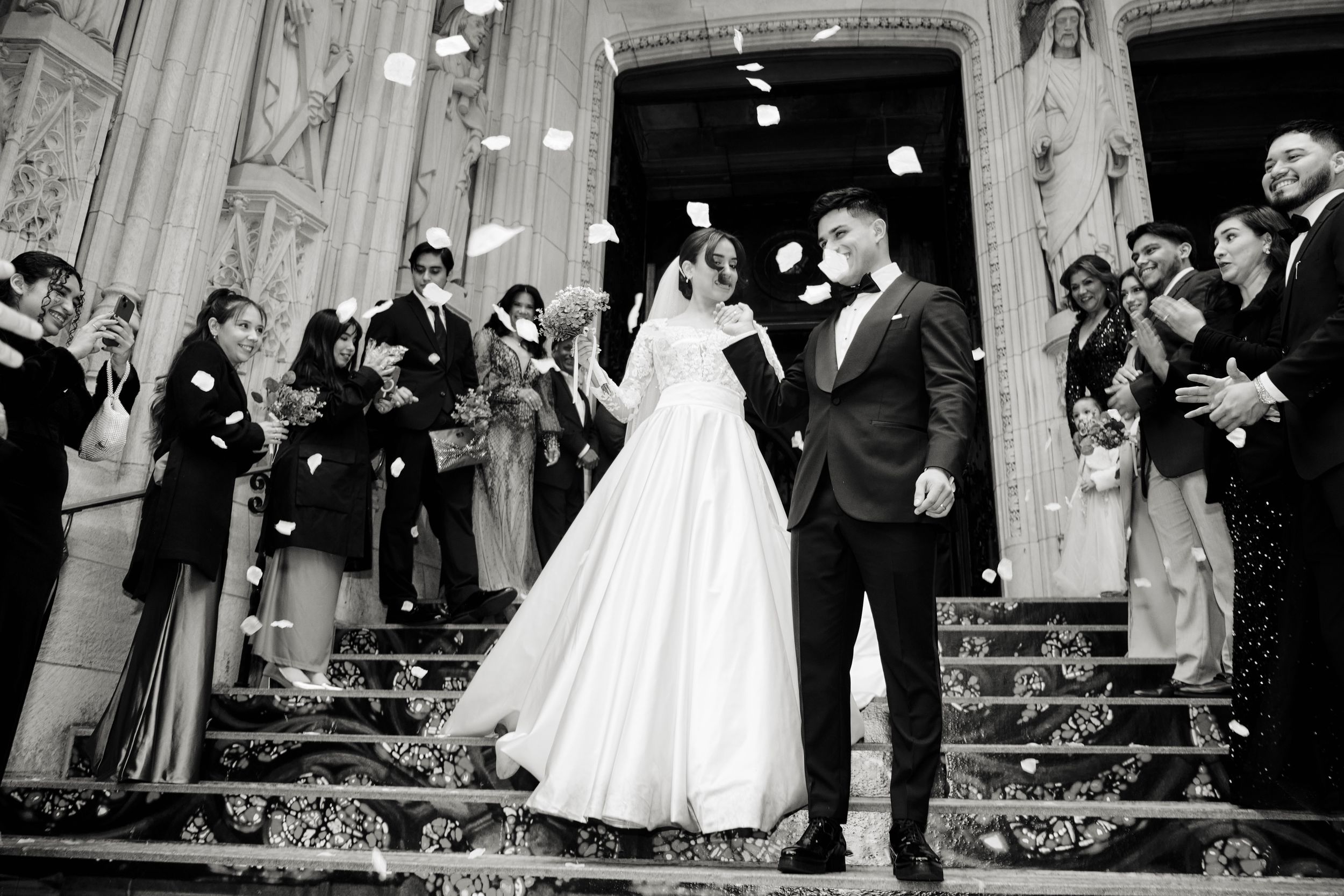 Newlyweds stand on ornate steps surrounded by guests throwing confetti in black and white photo.