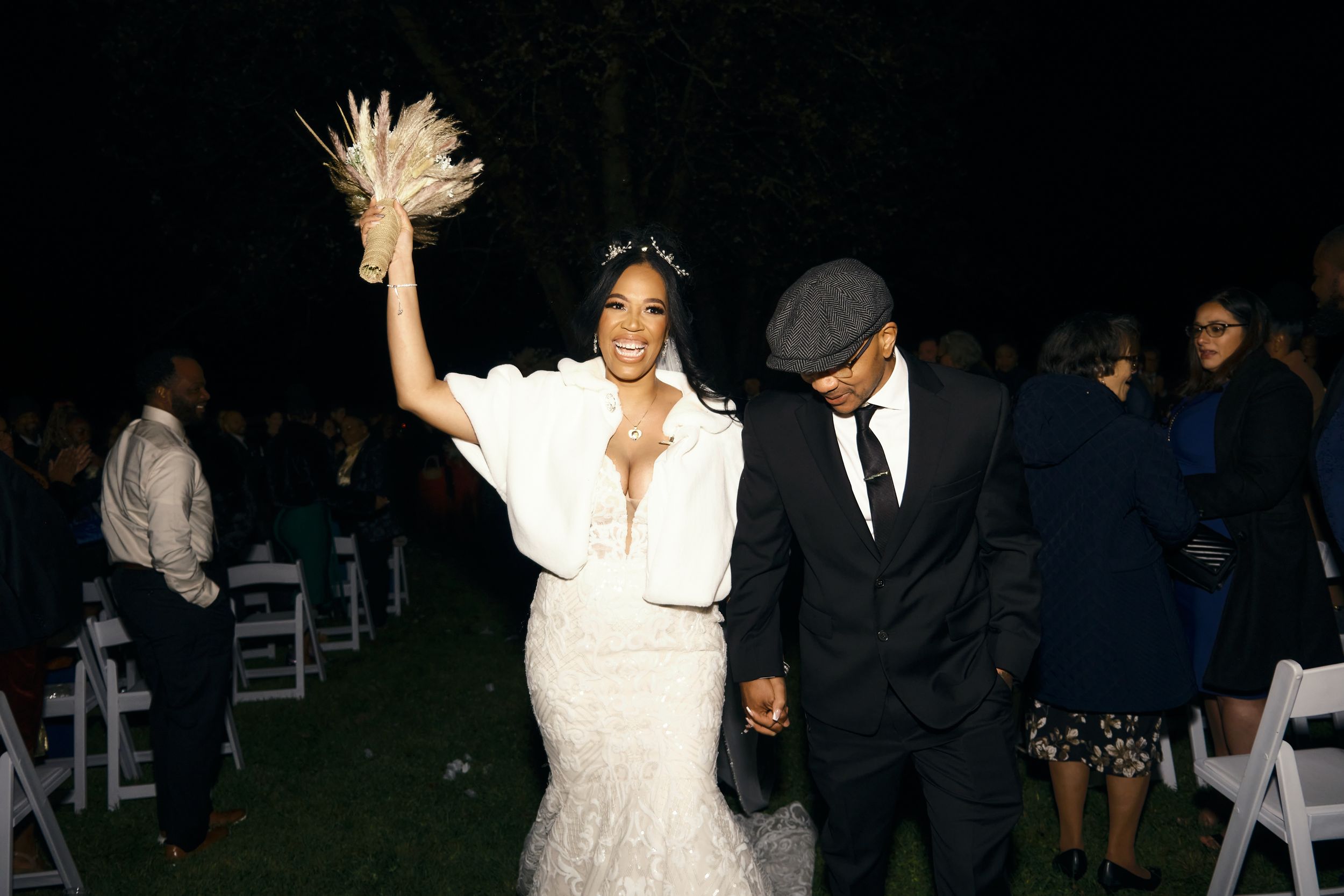 Celebratory moment at outdoor evening wedding as couple walks among white chairs.