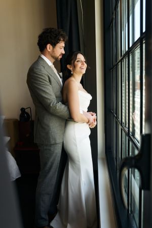 Couple in wedding attire shares intimate moment while looking through large window.