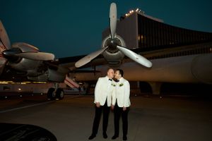 Couple in white outfits standing in front of vintage propeller aircraft at night.