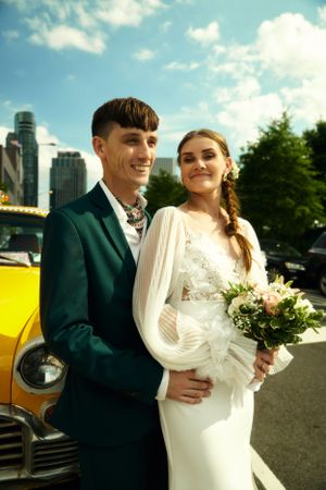 Wedding couple poses in front of yellow vintage car on urban street with modern buildings behind.