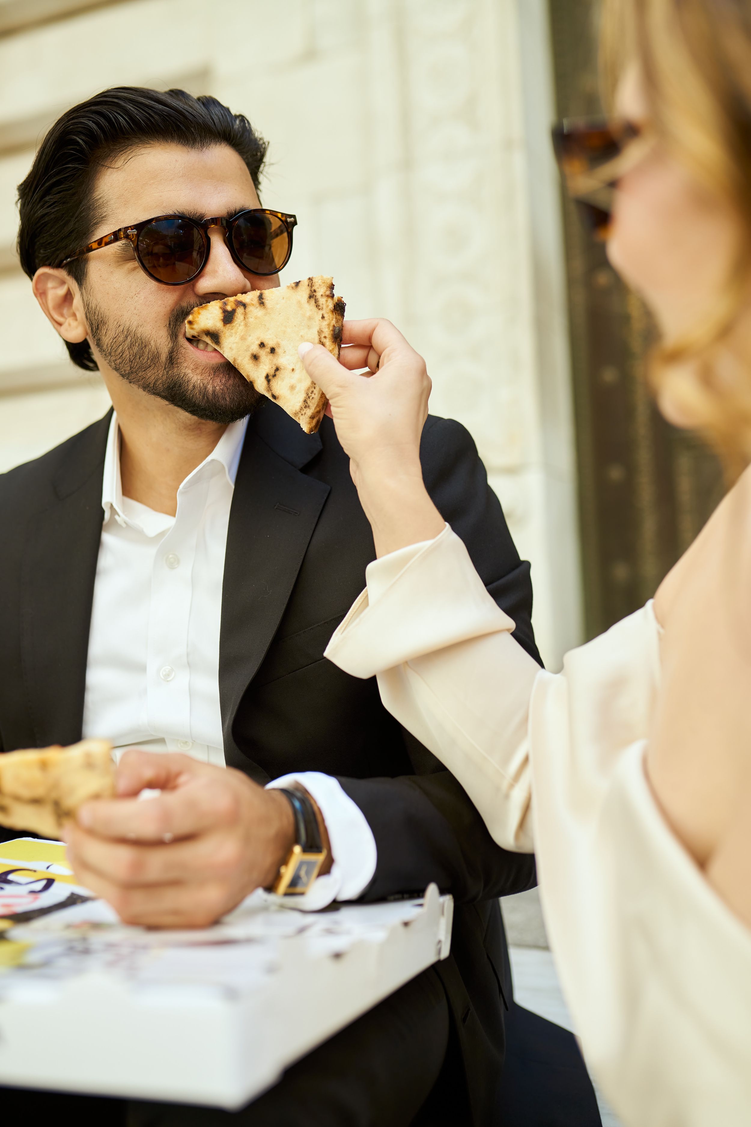 Couple shares pizza during casual moment in formal wear outside building.
