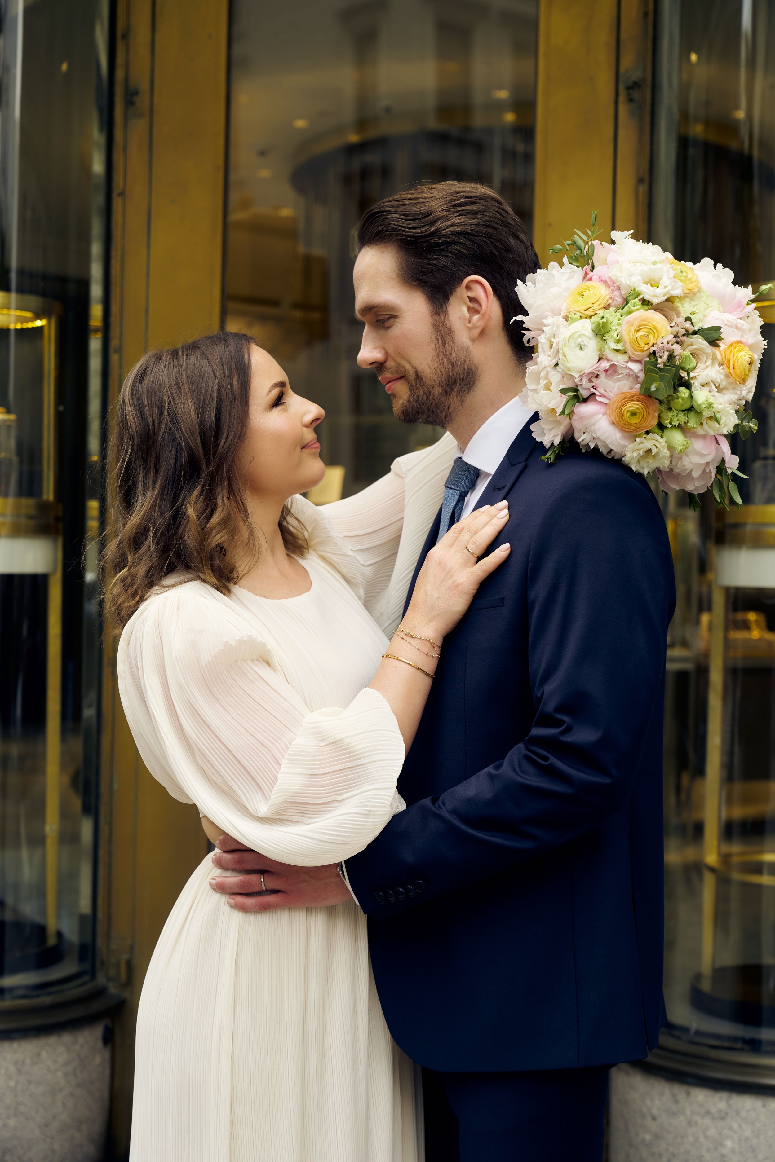 Romantic couple in wedding attire embrace outside revolving doors with spring bouquet.