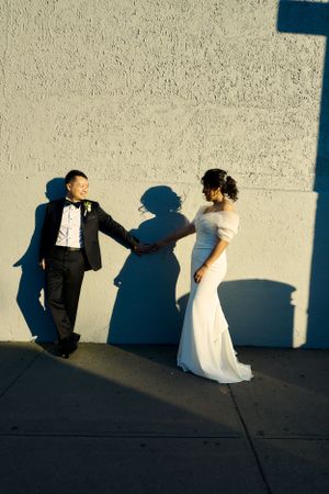 A couple in formal attire cast long shadows on a white wall during golden hour.