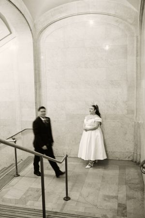 Black and white photo of couple in formal wear standing in arched marble hallway.