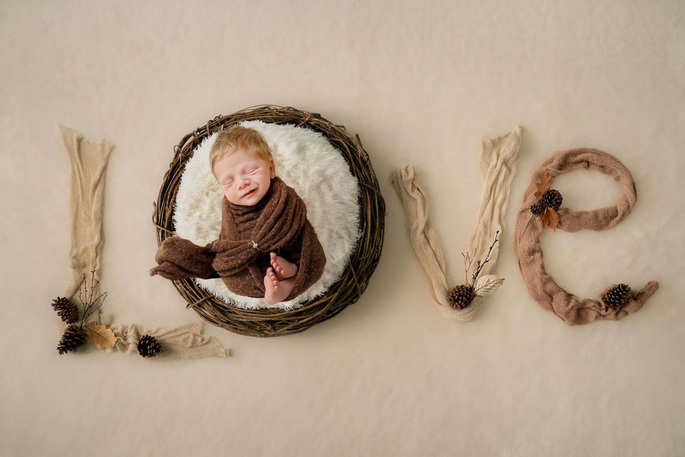 Sleeping newborn wrapped in brown swaddle posed in nest spelling LOVE with natural decor.