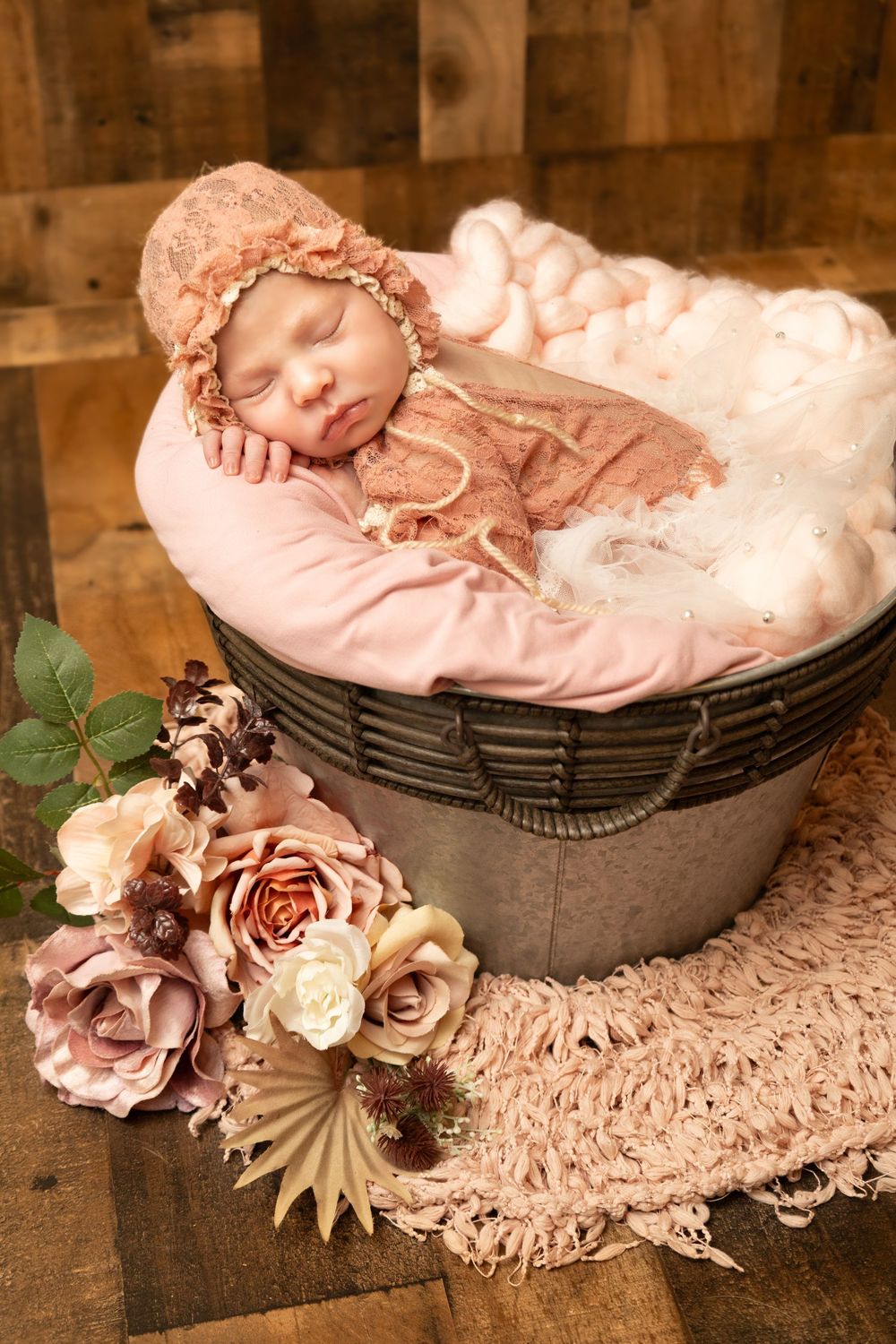Sleeping newborn in pink lace bonnet resting in metal bucket with floral arrangement on wood.