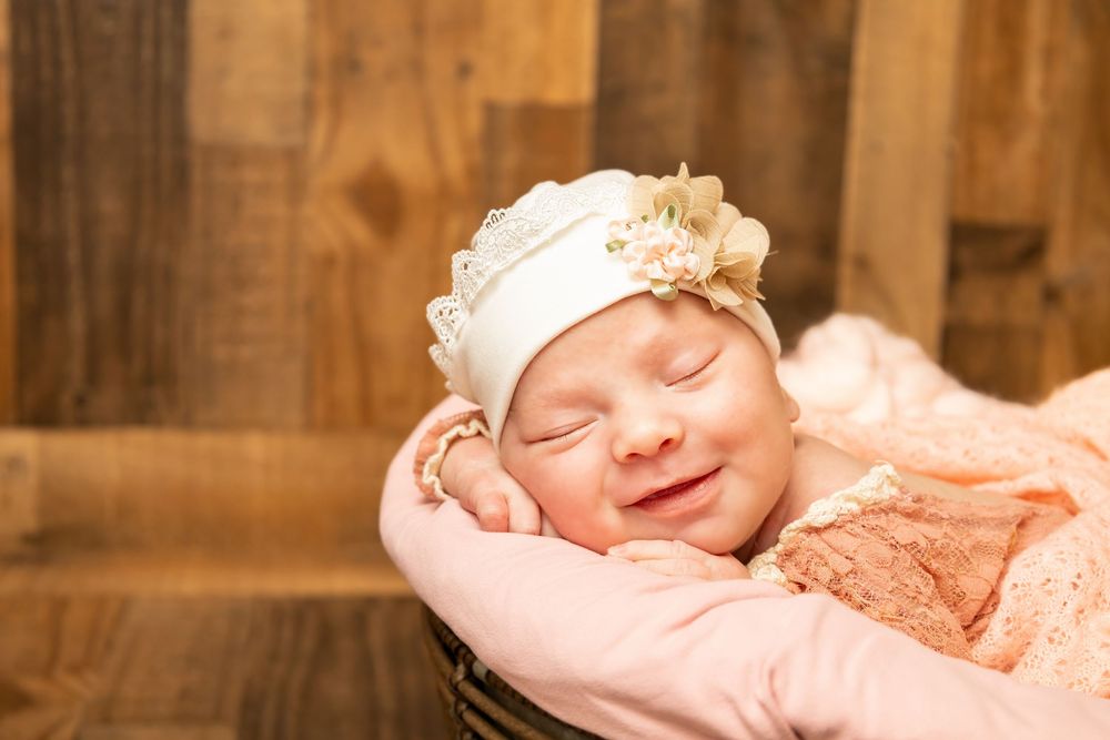 Smiling sleeping newborn in white floral hat nestled in pink blanket inside dark basket.