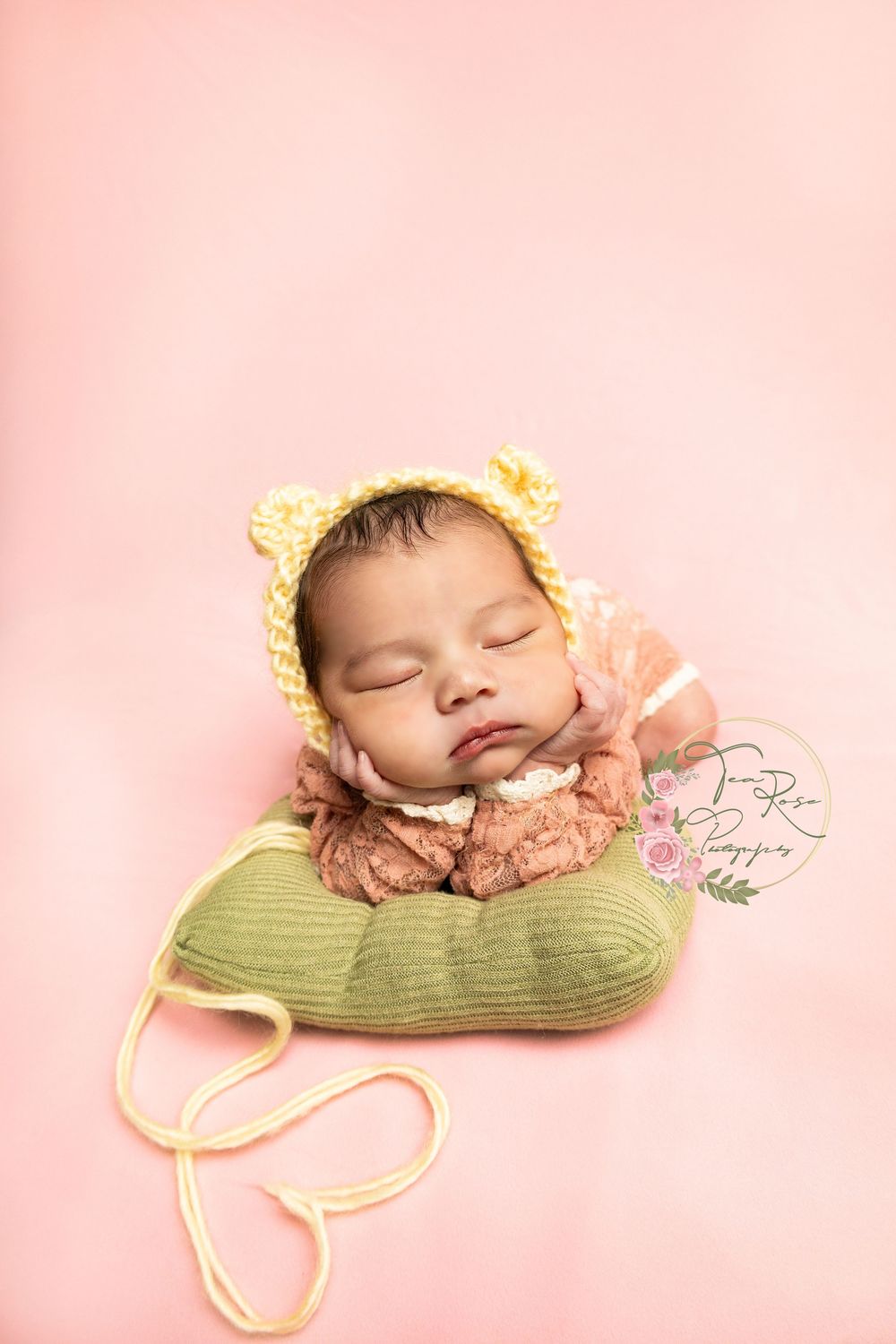 Sleeping newborn in yellow bear bonnet resting on green pillow against soft pink background.