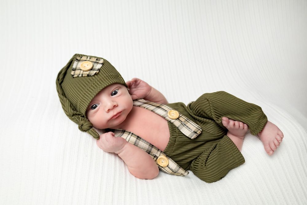 Alert newborn in olive green outfit with suspenders and matching hat on white blanket.