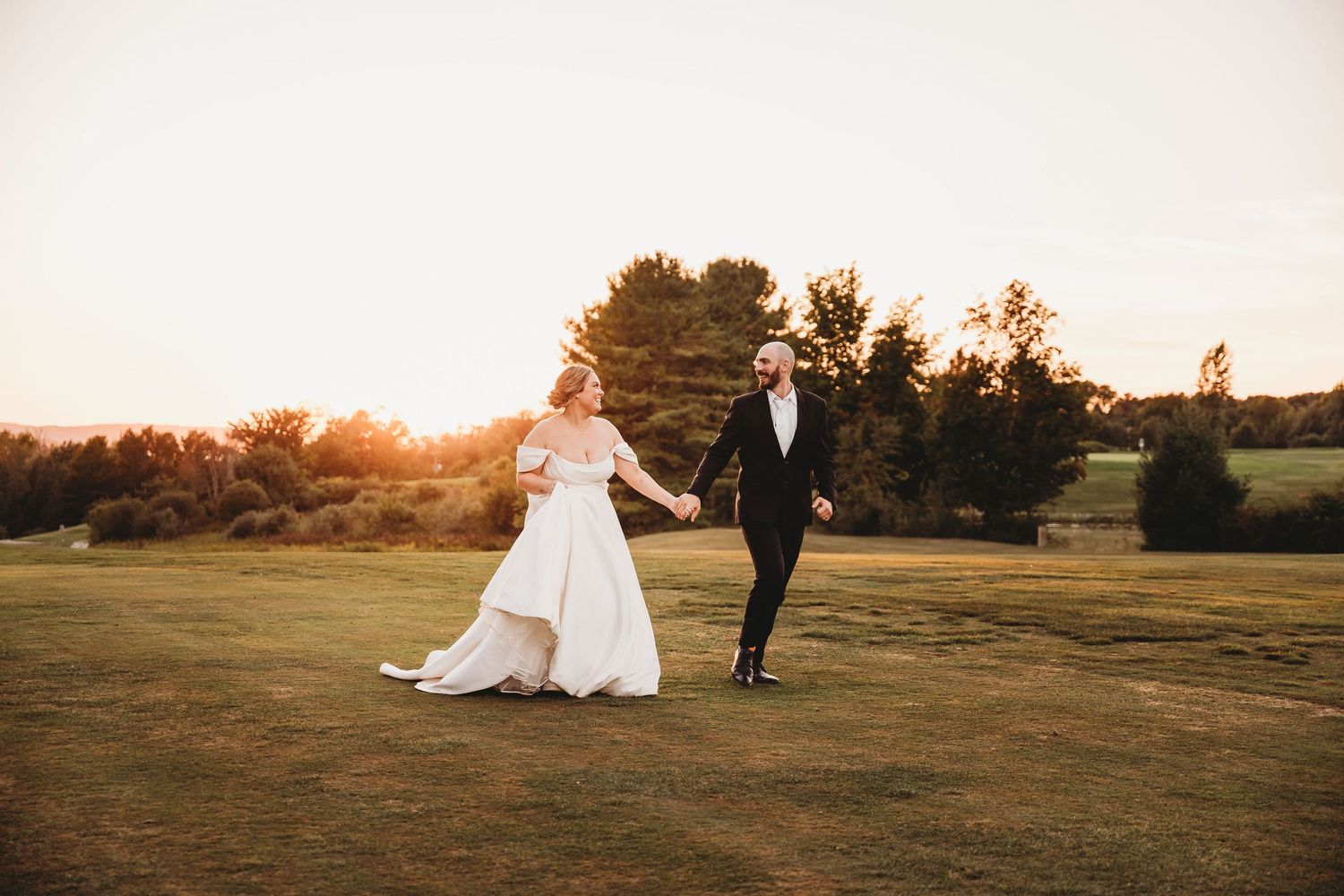 Golden sunset illuminates a couple in formal attire walking across an open field at dusk.