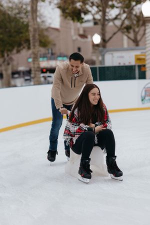 couple ice skating outdoors, with one partner pushing the other on a skate aid while smiling