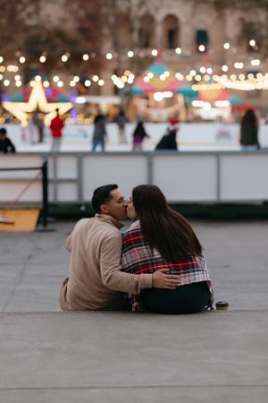 couple sitting on step near an ice skate rink, leaning in for a kiss with colorful lights and skaters blurred in the background
