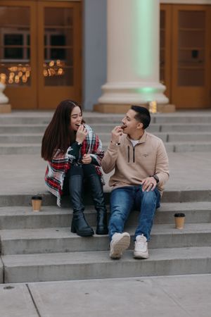 couple sitting on outdoor steps, laughing and sharing snacks and hot coco beside a building with columns