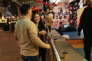 couple standing at a carnival game booth at night, smiling as the woman hold a plush prize with colorful lights and prizes behind them