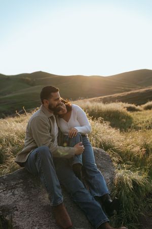 couple sitting together on a rock on a hillside overlooking hills at sunset