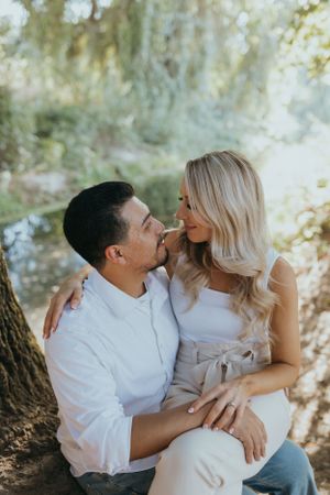 couple standing sitting closely together outdoors, smiling at each other while leaning against a tree in soft natural light