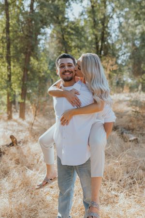 couple outdoors in a wooded area, with one partner giving the other a piggyback ride while smiling