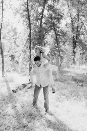 black and white image of a couple outdoors in a wooded area, one partner carrying the other on their back while walking