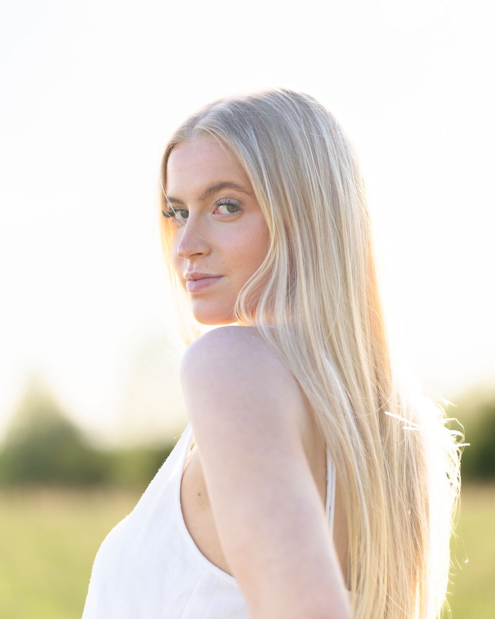 A person with long blonde hair poses in a white top against a sunset backdrop.