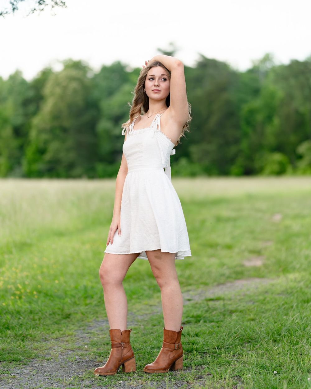 A person in a white dress and brown boots poses on a dirt path near green trees.