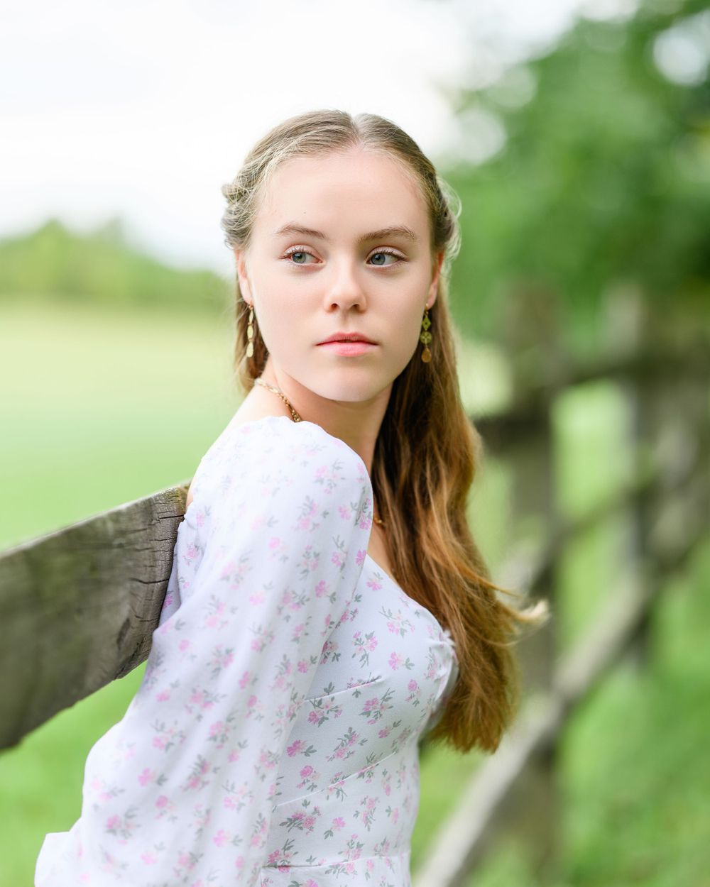 Outdoor portrait of someone in a floral dress leaning against wooden fence in natural setting.