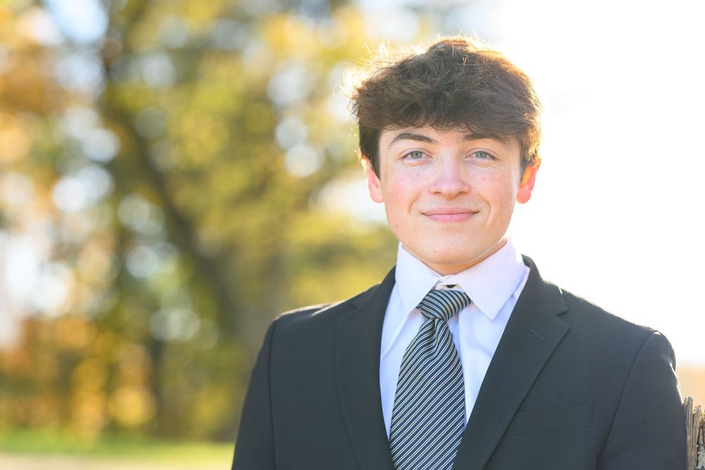 Professional headshot outdoors featuring someone in a dark suit and striped tie with autumn trees behind.