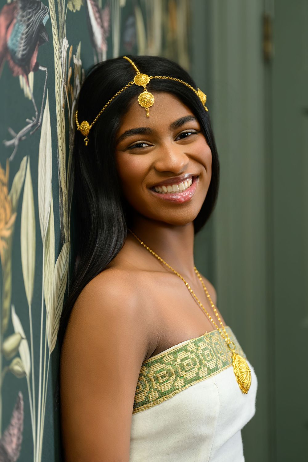 Smiling young woman in white and gold traditional attire with gold headpiece, posing against a floral wallpaper backdrop.