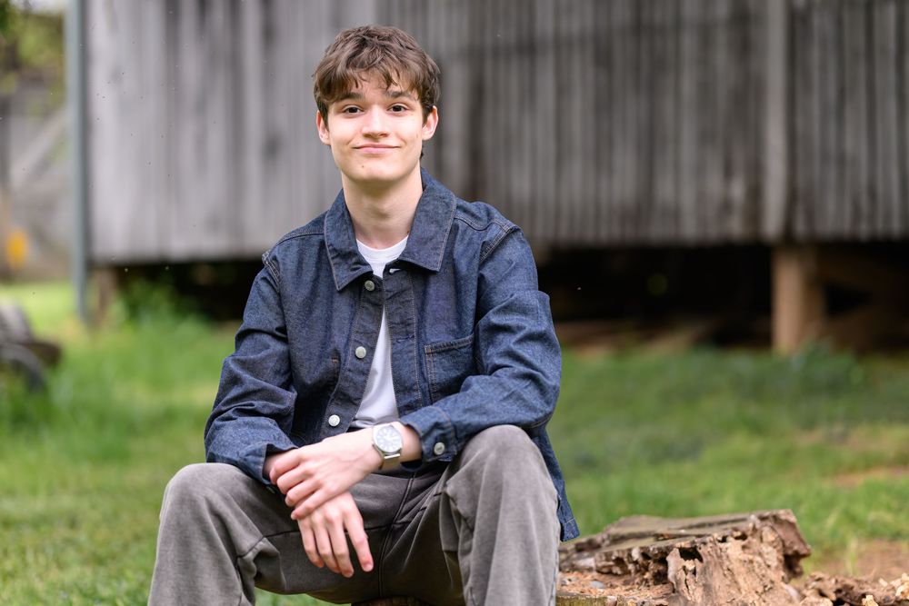 A young man in a denim jacket sits outdoors on a log, smiling casually in a green backyard setting.