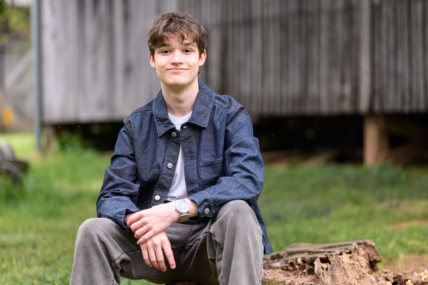 A young man in a denim jacket sits outdoors on a log, smiling casually in a green backyard setting.