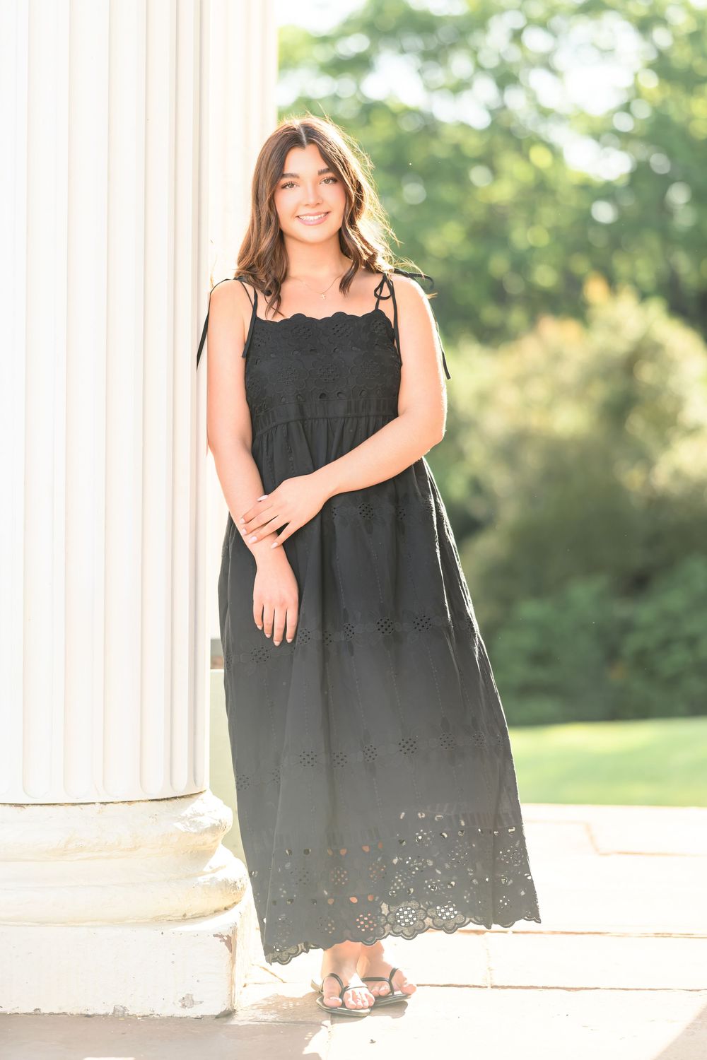 Young woman smiling in black eyelet maxi dress, posing by white column outdoors in lush green setting.