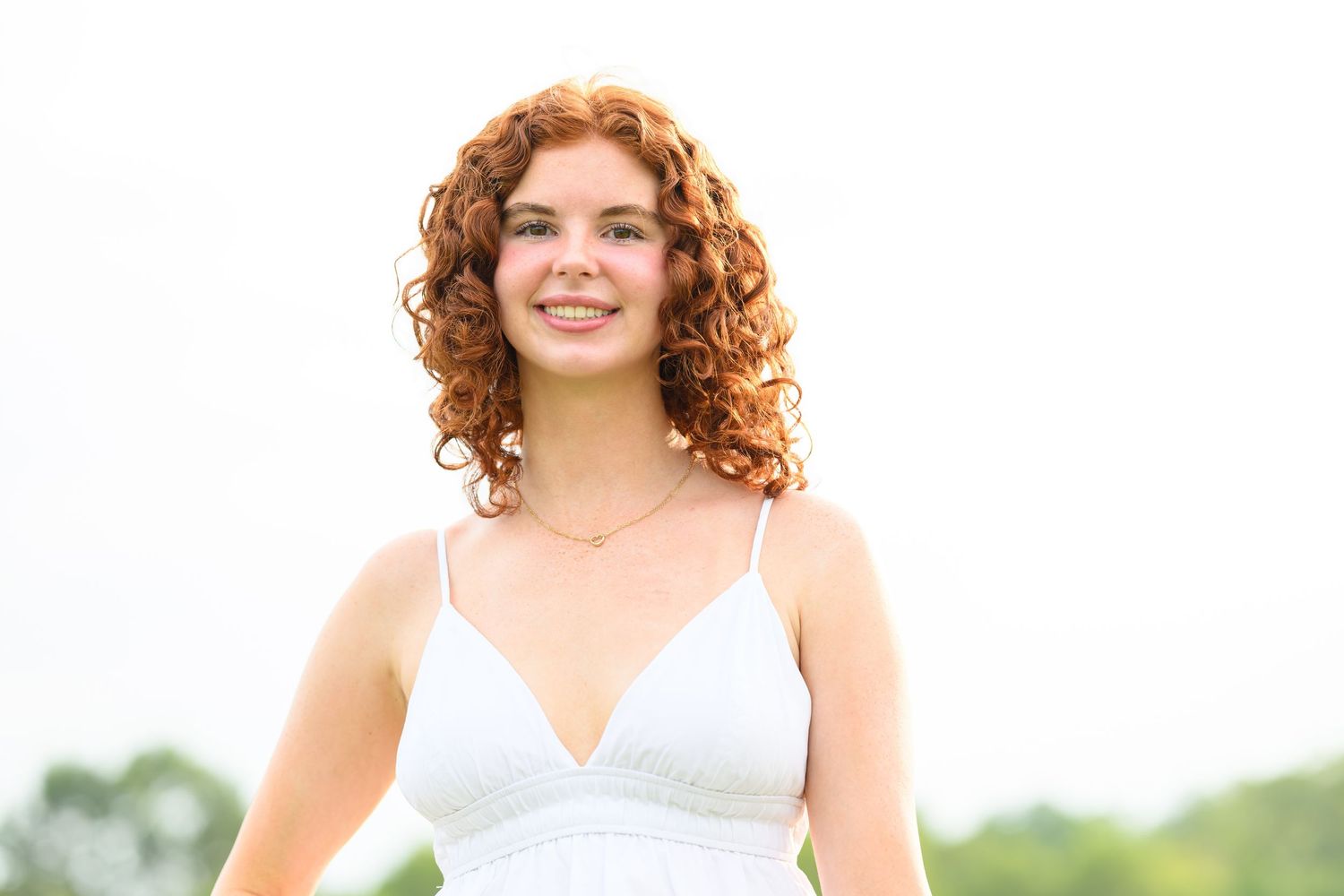 Young woman with curly red hair smiling outdoors in a white dress on a bright day.