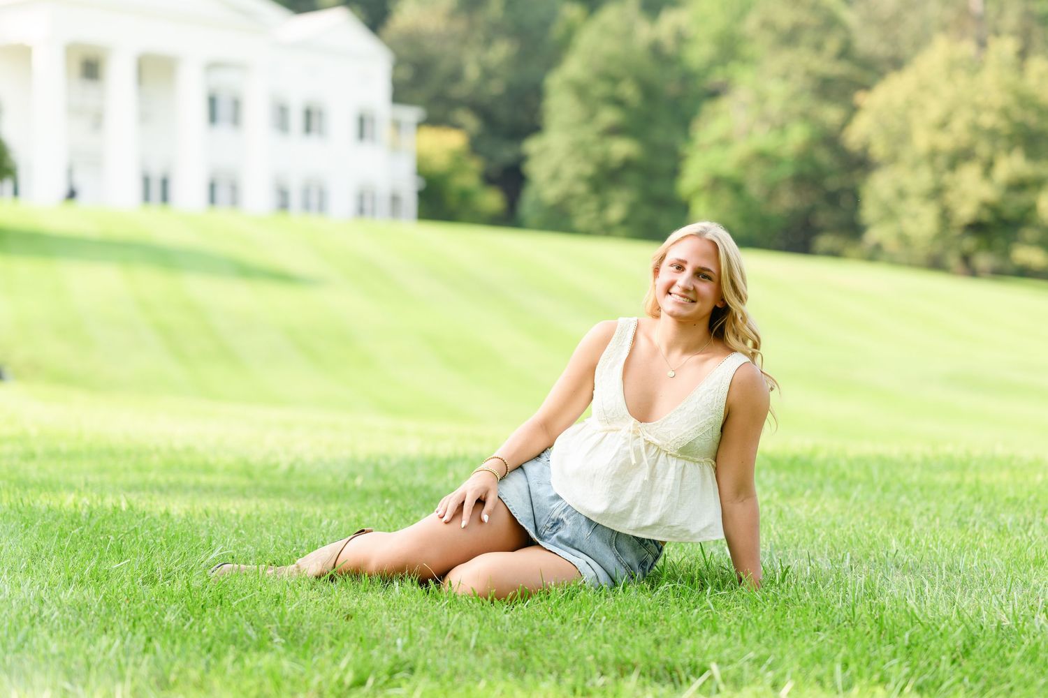 Young blonde woman sitting on lush green grass outdoors, wearing white top and denim shorts, with white mansion in background.