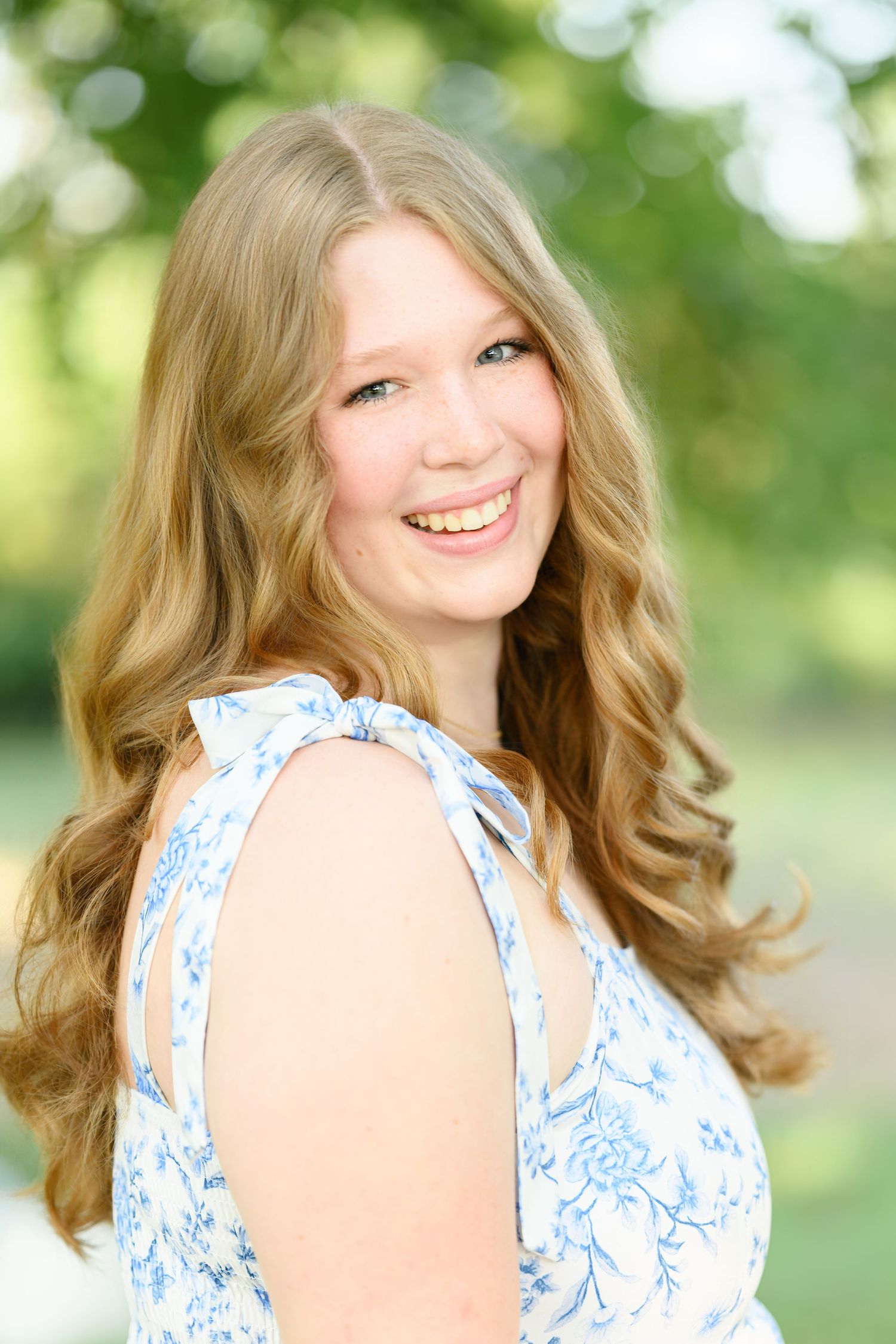 Smiling young woman with long blonde wavy hair wearing a blue floral dress outdoors in a green park setting.