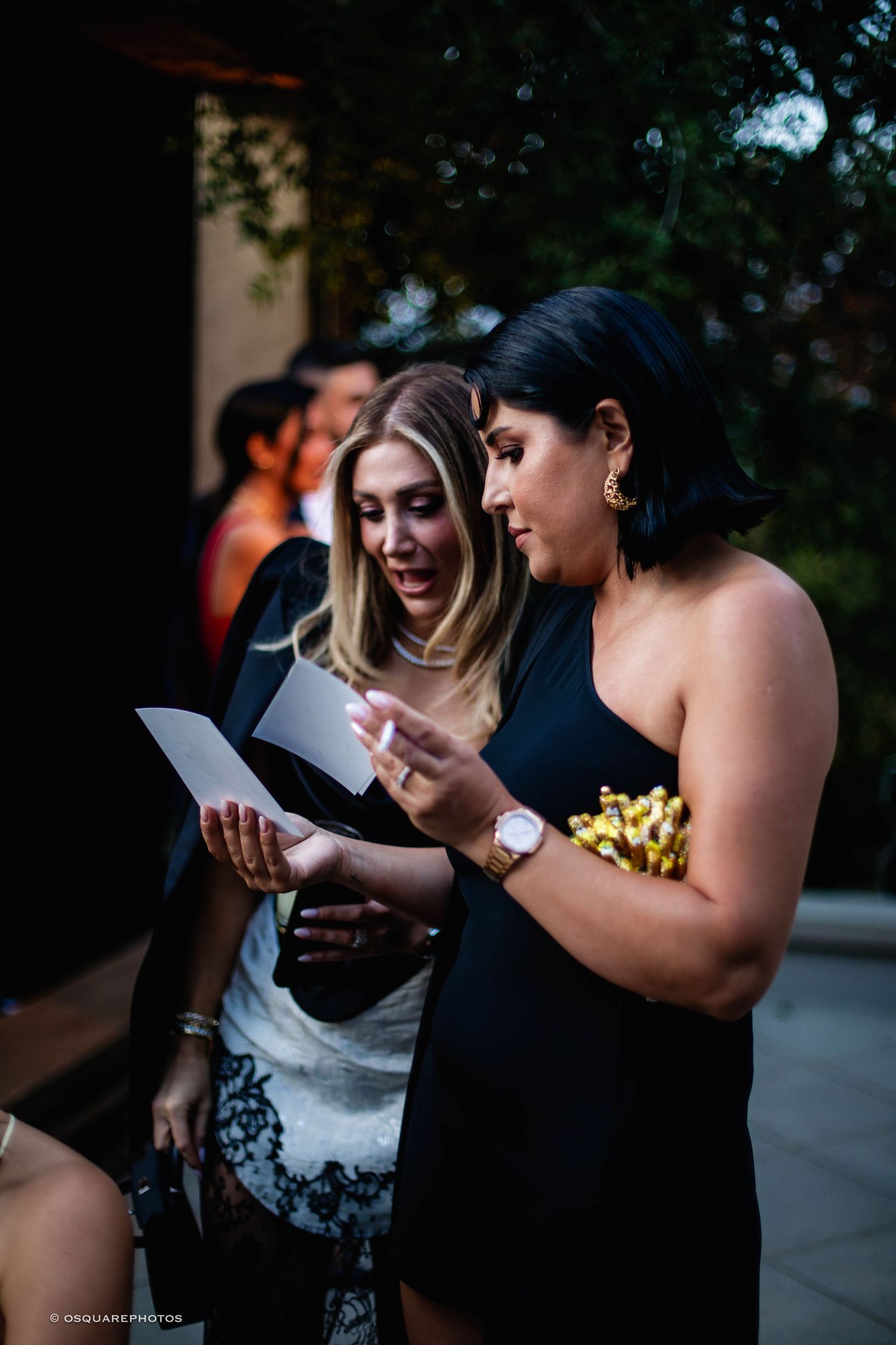 Two women in formal attire share a joyful moment while reading papers together outdoors.