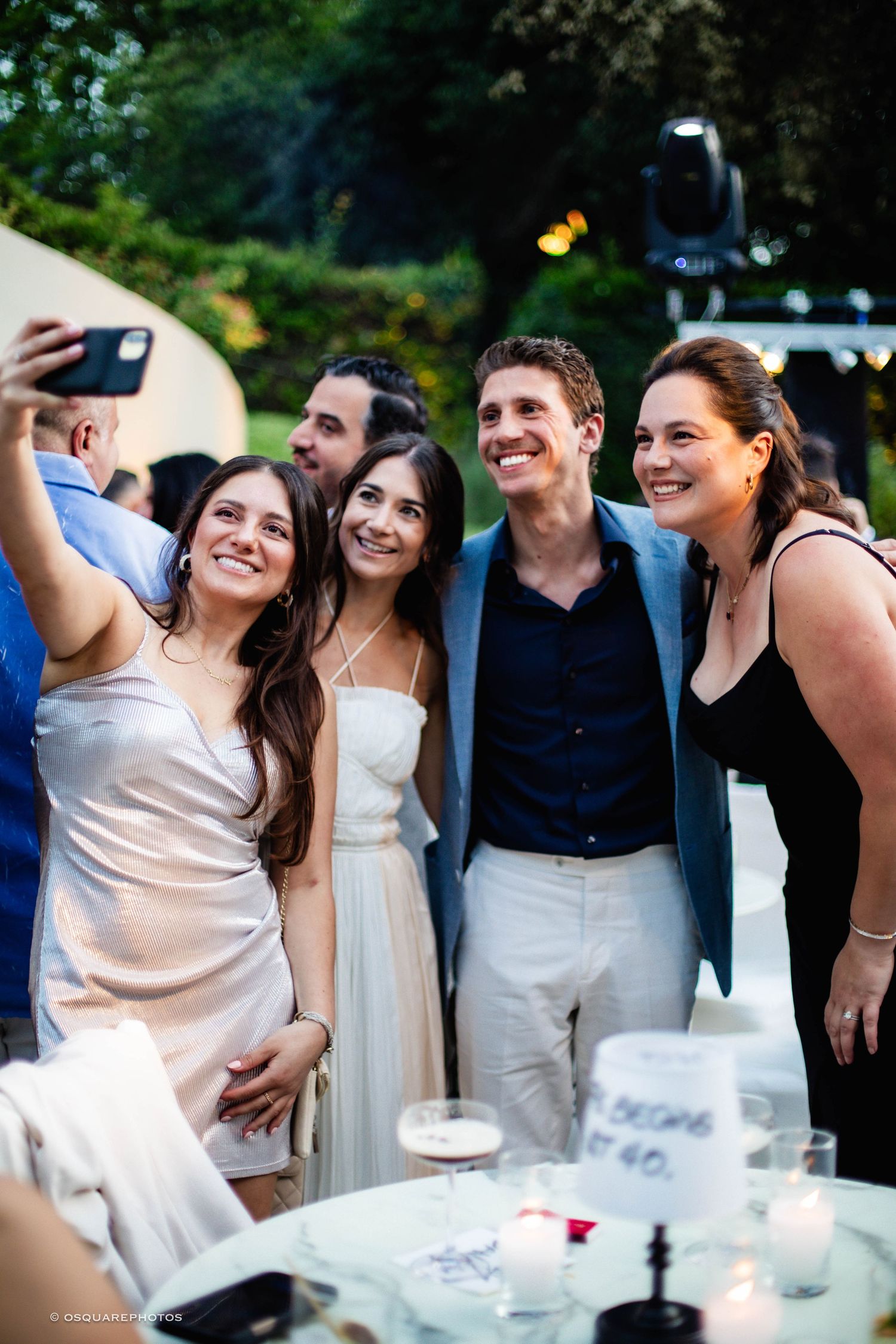 Wedding party guests smile and take selfies during an outdoor reception gathering.