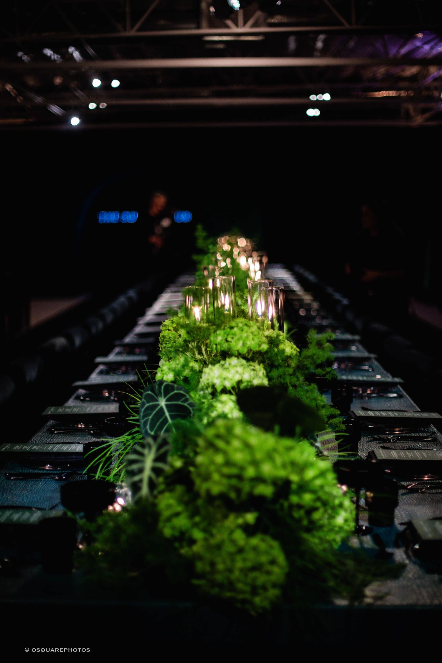Dramatic lighting illuminates green floral centerpieces on dark tables at an elegant event setup.
