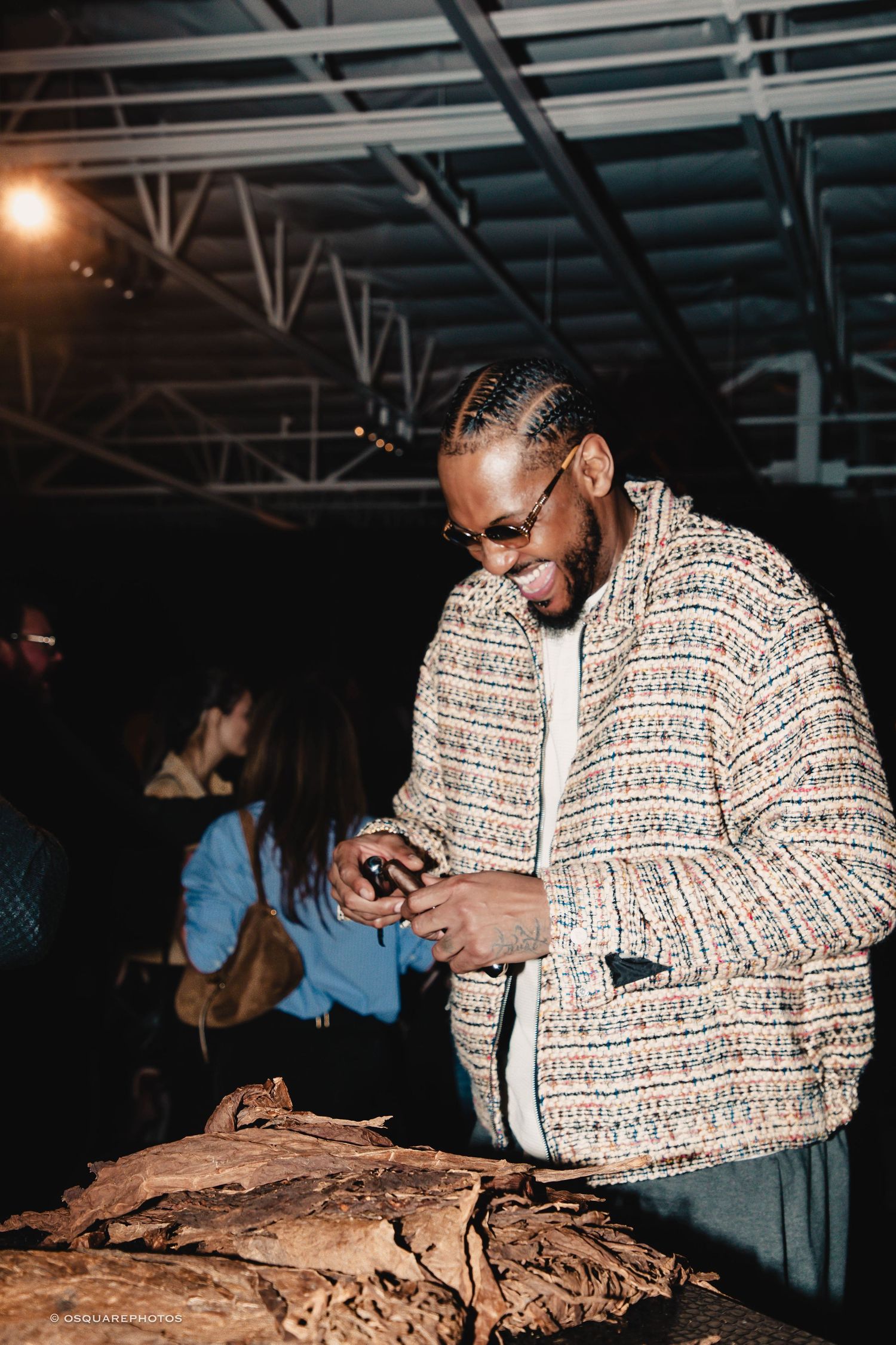 A person wearing a patterned jacket examines and sorts tobacco leaves on a wooden surface in a dark warehouse setting.
