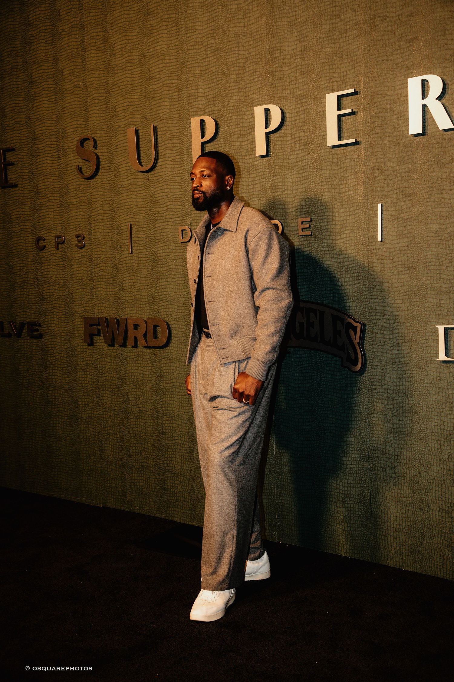 A person in a light-colored outfit stands against a textured wall with dramatic shadows and lighting effects.