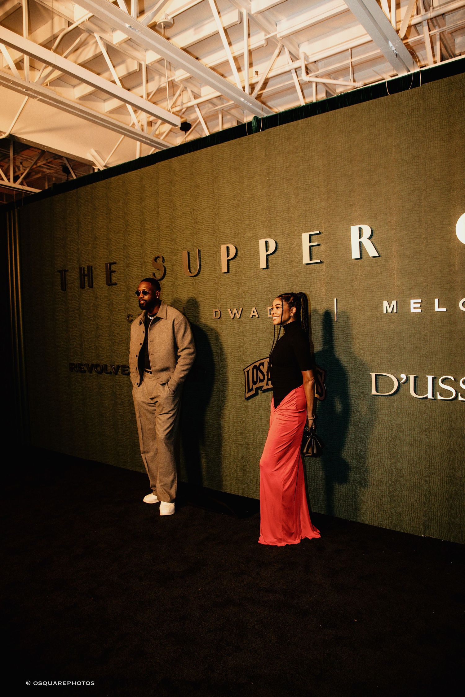 Two people stand against a dark wall with 'THE UPPER' signage in an industrial-style space with exposed ceiling beams.