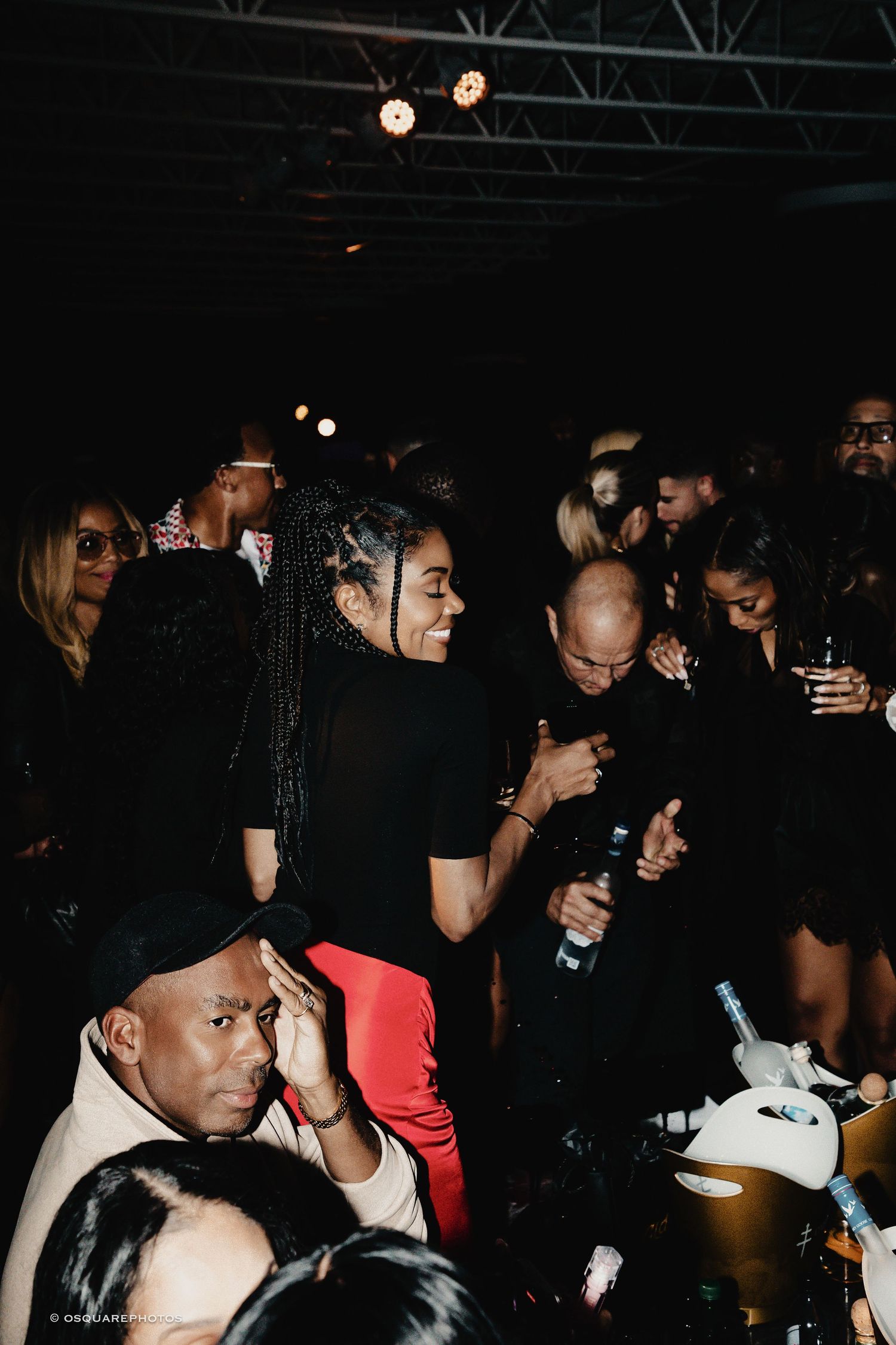 Dark nightclub scene showing people socializing and dancing in a dimly lit space with spotlights overhead.