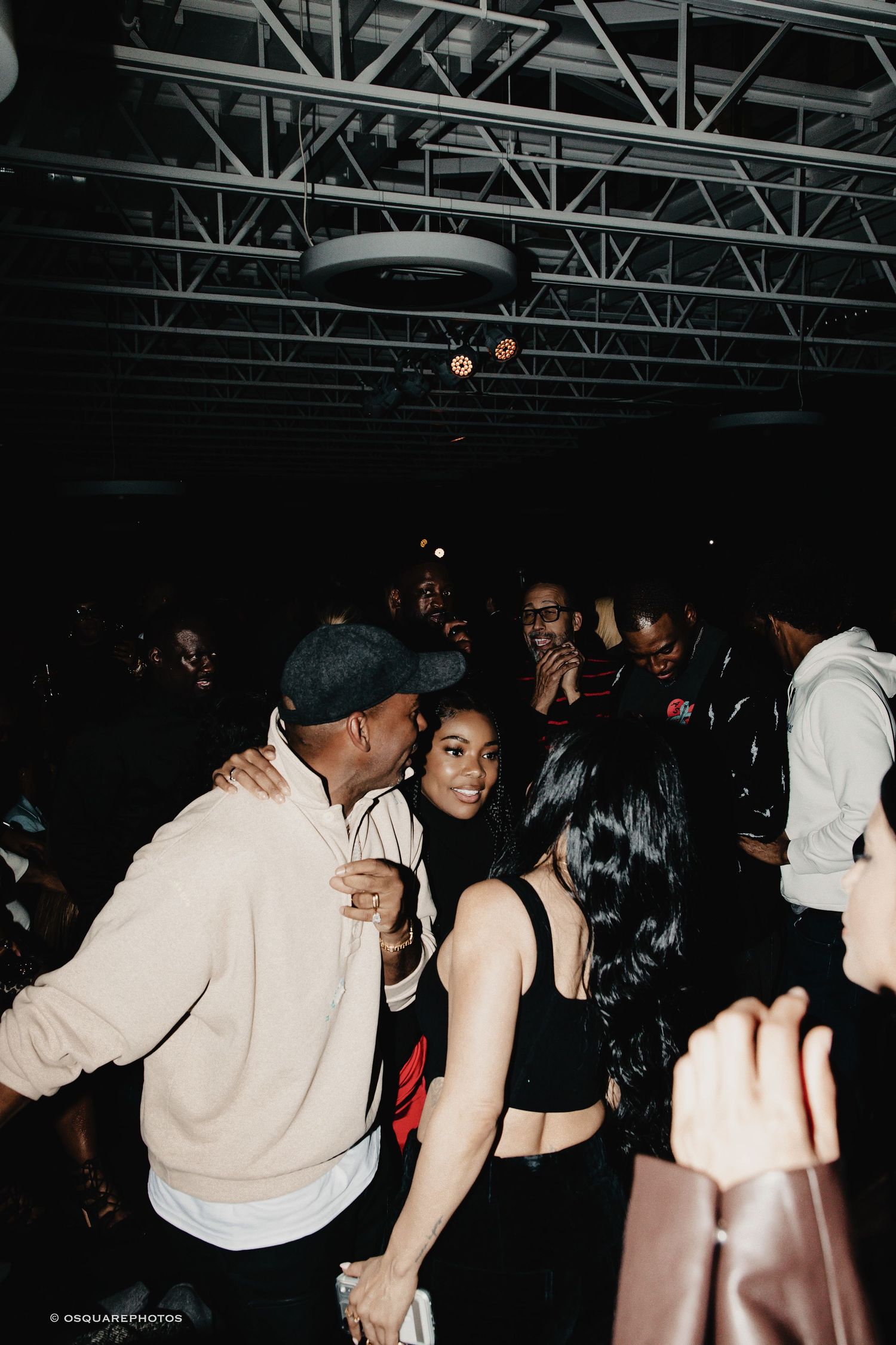 People dancing and socializing in a dimly lit nightclub with industrial ceiling beams visible above.