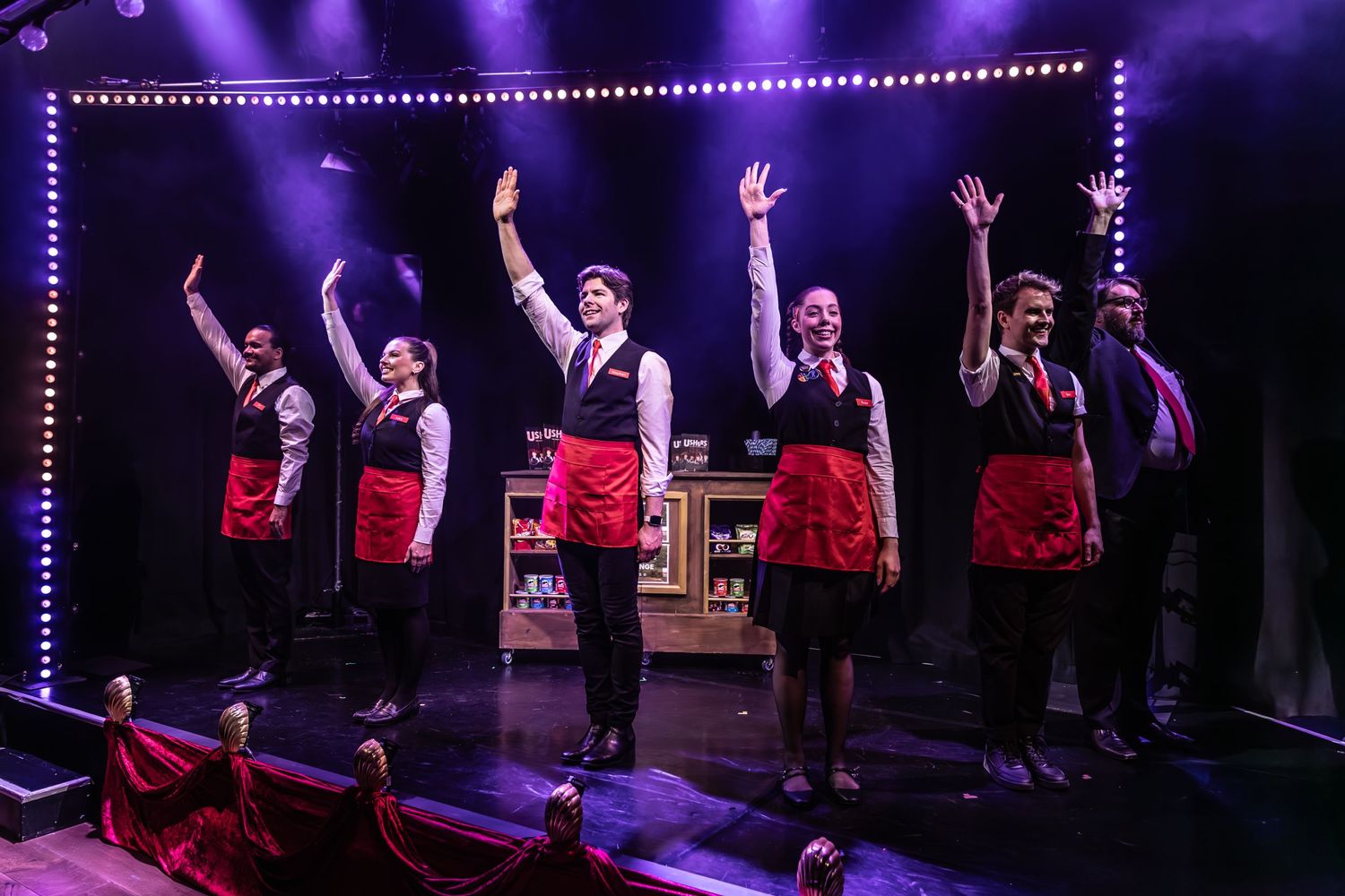 Theater ensemble performs in matching red and black costumes under purple stage lighting.