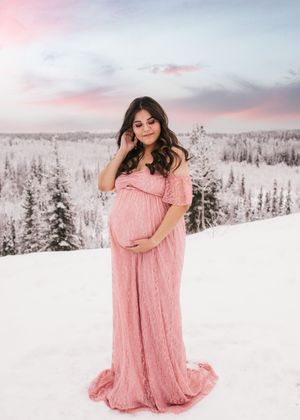 Pregnant woman wearing a flowing pink dress standing in a snowy winter landscape during a maternity session