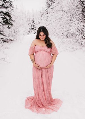 Pregnant woman in a flowing pink dress standing in a snowy forest during a maternity session