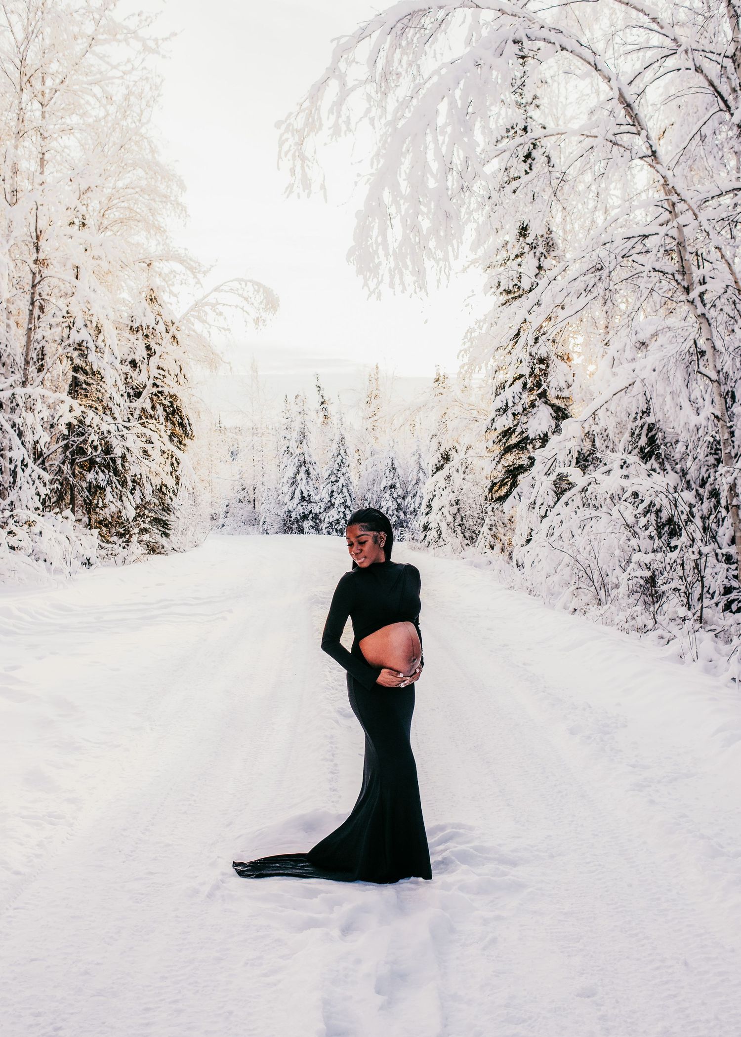Pregnant woman wearing a long black dress standing in a snowy winter forest during a maternity session