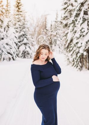 Pregnant woman wearing a blue dress standing in a snowy winter forest during a maternity session