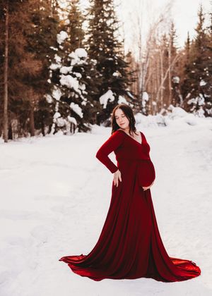 Pregnant woman wearing a flowing red dress standing in a snowy forest during a maternity session