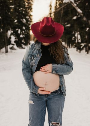Pregnant woman wearing a red hat and denim jacket standing outdoors in winter during a maternity session